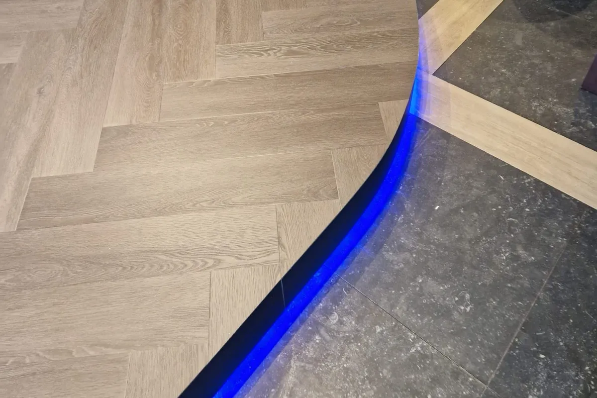 Gebogen visgraat-inleg in PVC met een blauwe LED-lichtrand, overgangsprofiel naar buitentegels op een terras.